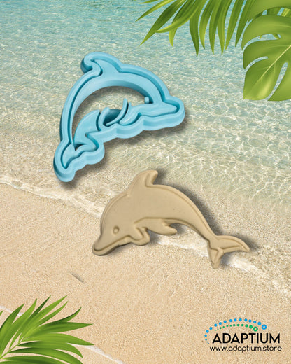 Blue dolphin polymer clay cutter with matching tan clay dolphin piece displayed on a sandy beach background with turquoise ocean waves and tropical leaves.